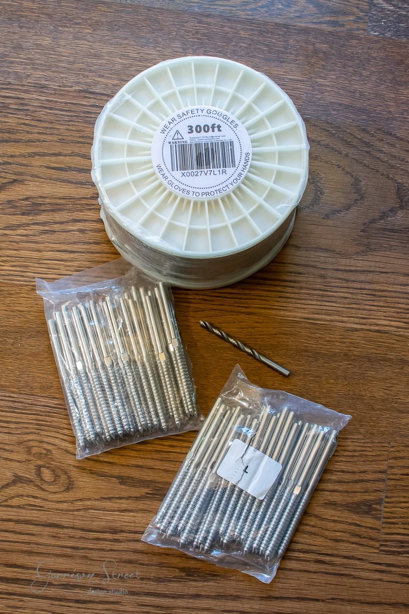 Roll of white thread labeled "300 ft," alongside two bags of metal nails and a single drill bit, sits on a wooden surface, perfect for your next DIY project. The label includes a reminder to wear safety goggles when crafting that cable railing masterpiece.