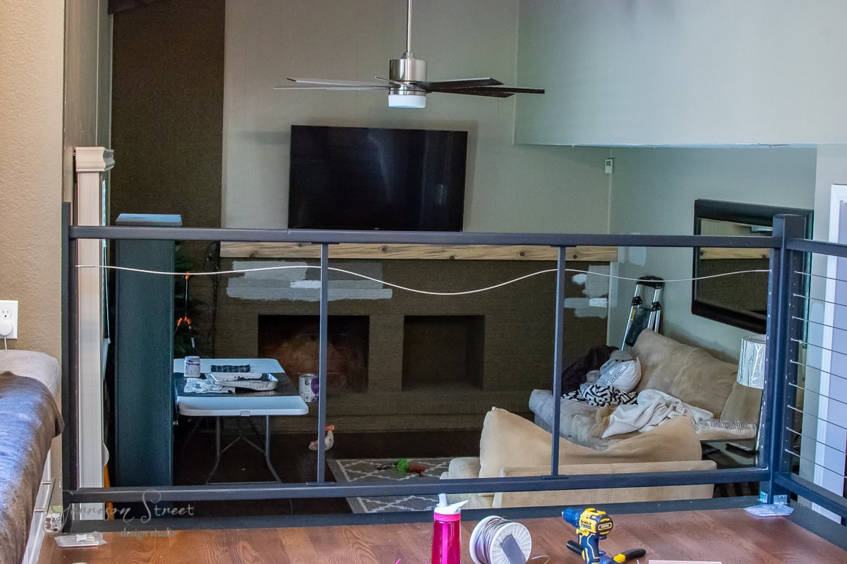 A living room under renovation features a modern ceiling fan, wall-mounted TV, and fireplace. A beige couch is partially covered with tools. A cable railing divides the foreground, adding to the DIY vibe, while supplies are scattered on a nearby table.