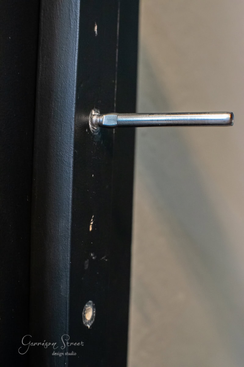 Close-up of a metal rod, reminiscent of a cable railing, protruding from a dark-colored door frame, with a small circular hole beneath it. The name "Garrison Street Design Studio" is faintly visible in the corner of the image, evoking a sleek DIY aesthetic.