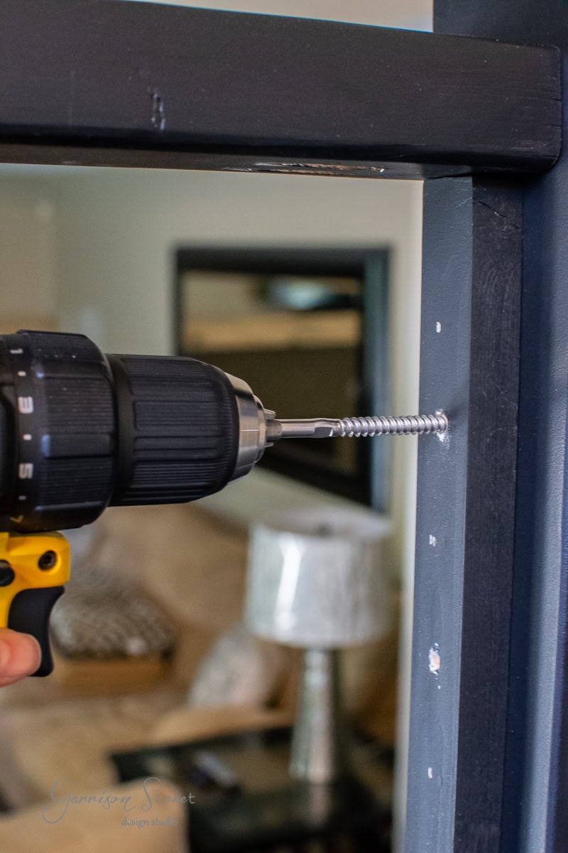 A hand expertly uses a black and yellow power drill to drive a screw into a dark painted wooden surface, part of the DIY cable railing project, with a blurred living room in the background.