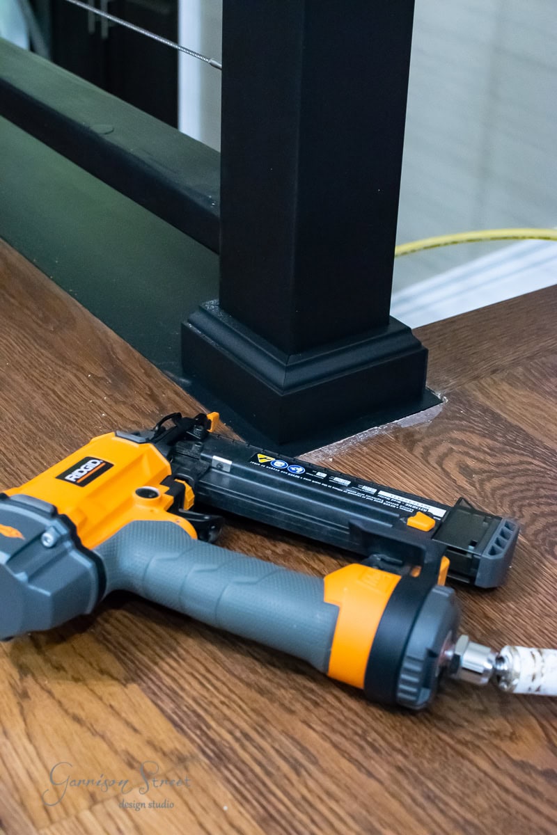 An orange and gray nail gun rests on a wooden floor beside a black post with cable railings, perfect for your next DIY project. A yellow power cord snakes across the scene, hinting at hands-on creativity in progress.