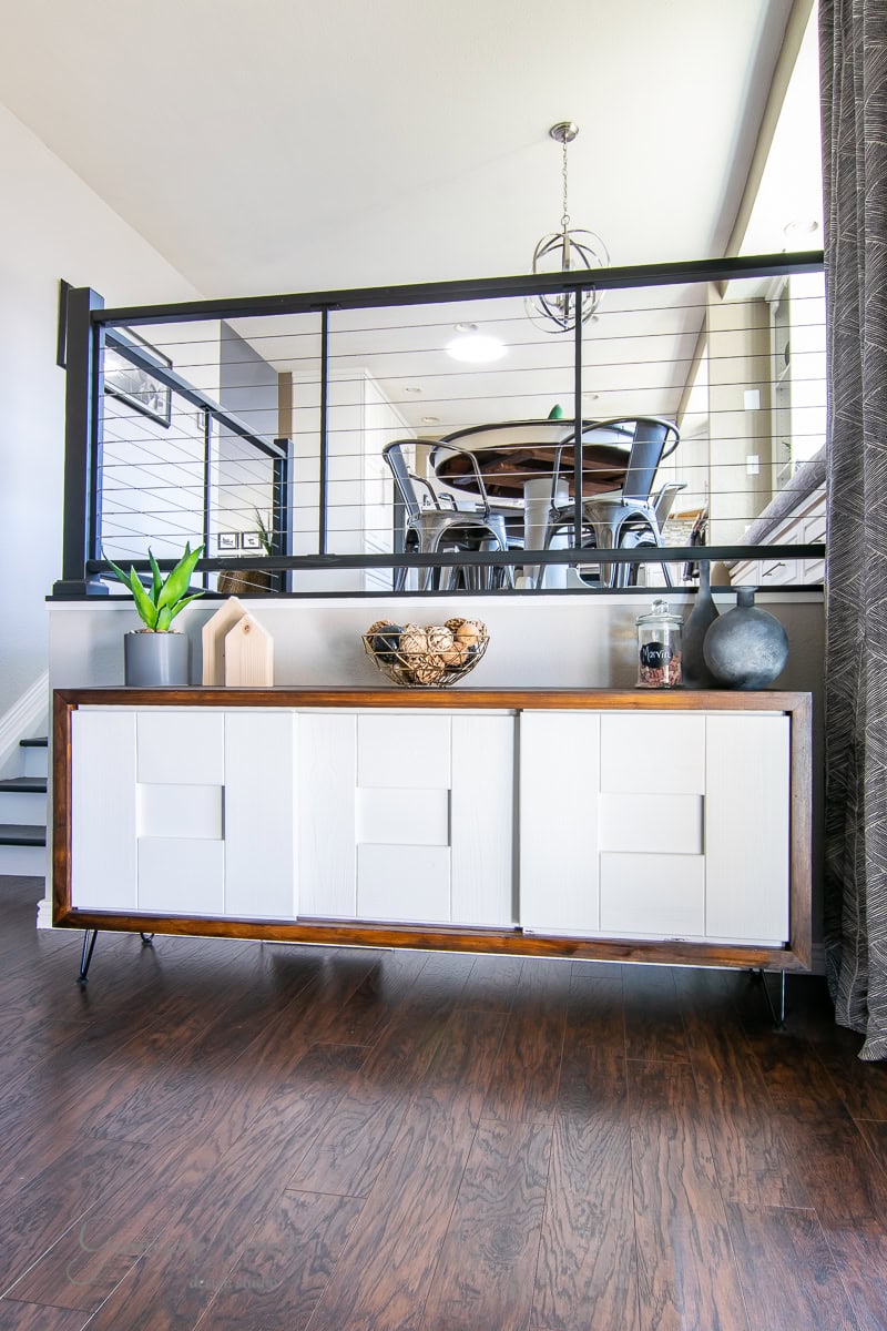 A modern living space features a white sideboard adorned with decorative items, including plants and a vase. The room boasts wooden floors and a sleek cable railing, hinting at DIY charm, with a dining table visible in the background.
