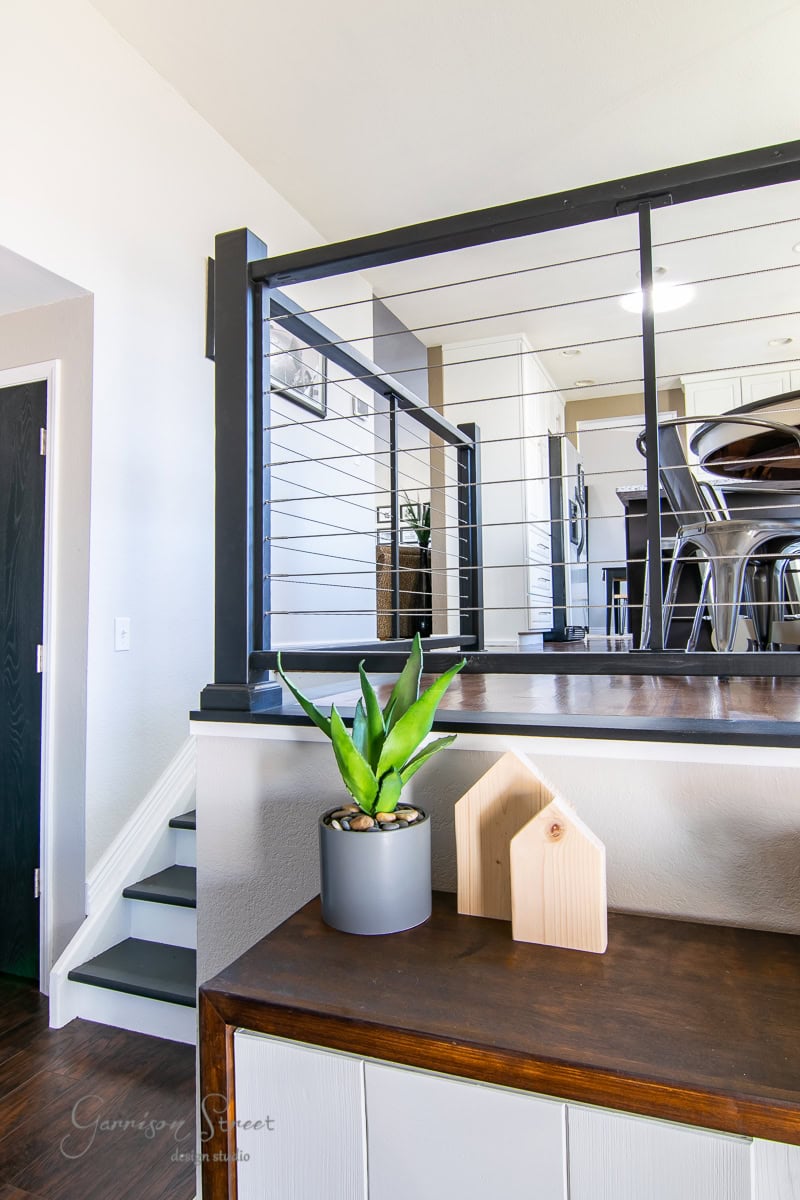 A modern interior showcases a wooden sideboard adorned with a green potted plant and wooden house-shaped decor. DIY cable railing and stairs guide the way to a dining area featuring a wooden table and metal chairs in the background.
