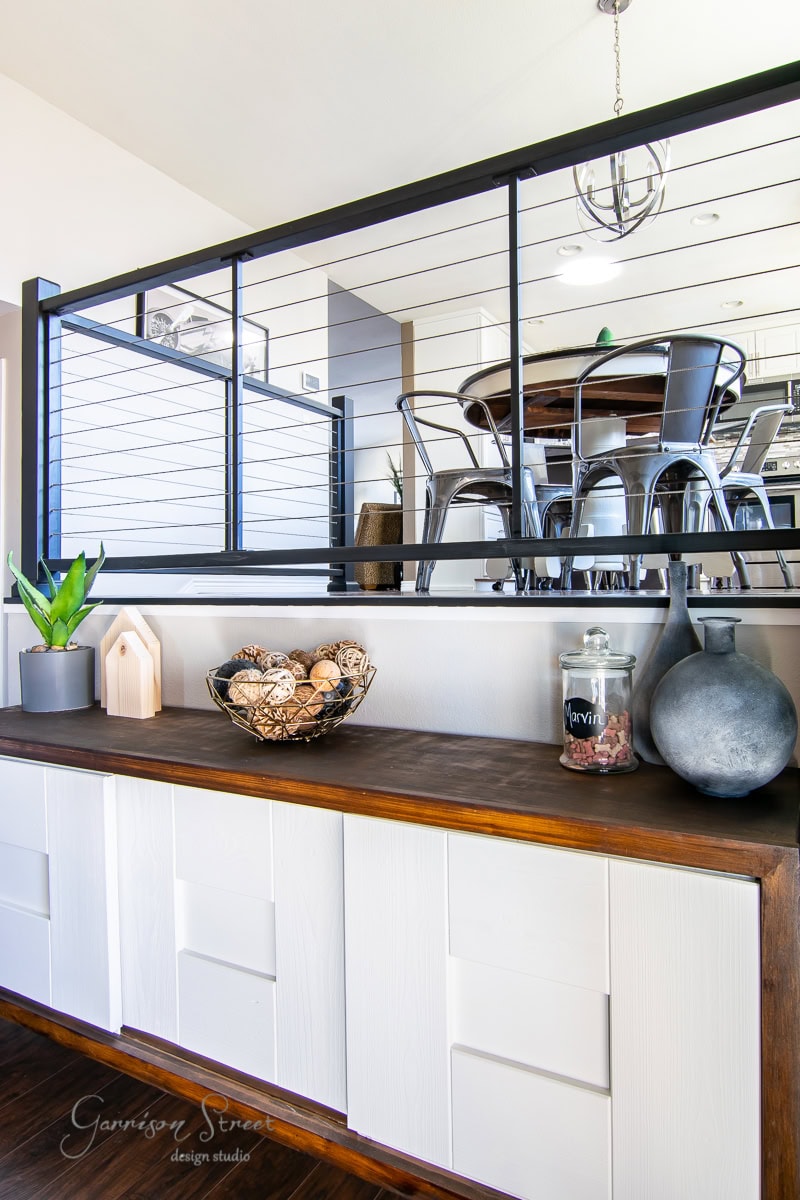 Modern interior with a wooden countertop showcasing DIY decor: a plant, a basket of balls, a large vase, and a jar labeled "penny." Above is a cable railing that artfully separates the space from the dining area with sleek metal chairs.