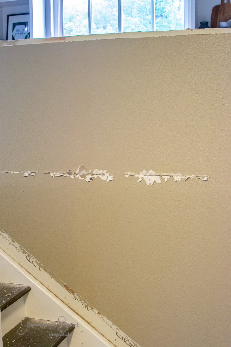A beige interior wall displays a long horizontal patch of chipped paint, revealing white underneath. The scene features a staircase with a sleek cable railing and a wooden banister, while a window above lets in natural light, offering the perfect opportunity for DIY enthusiasts to refresh their space.
