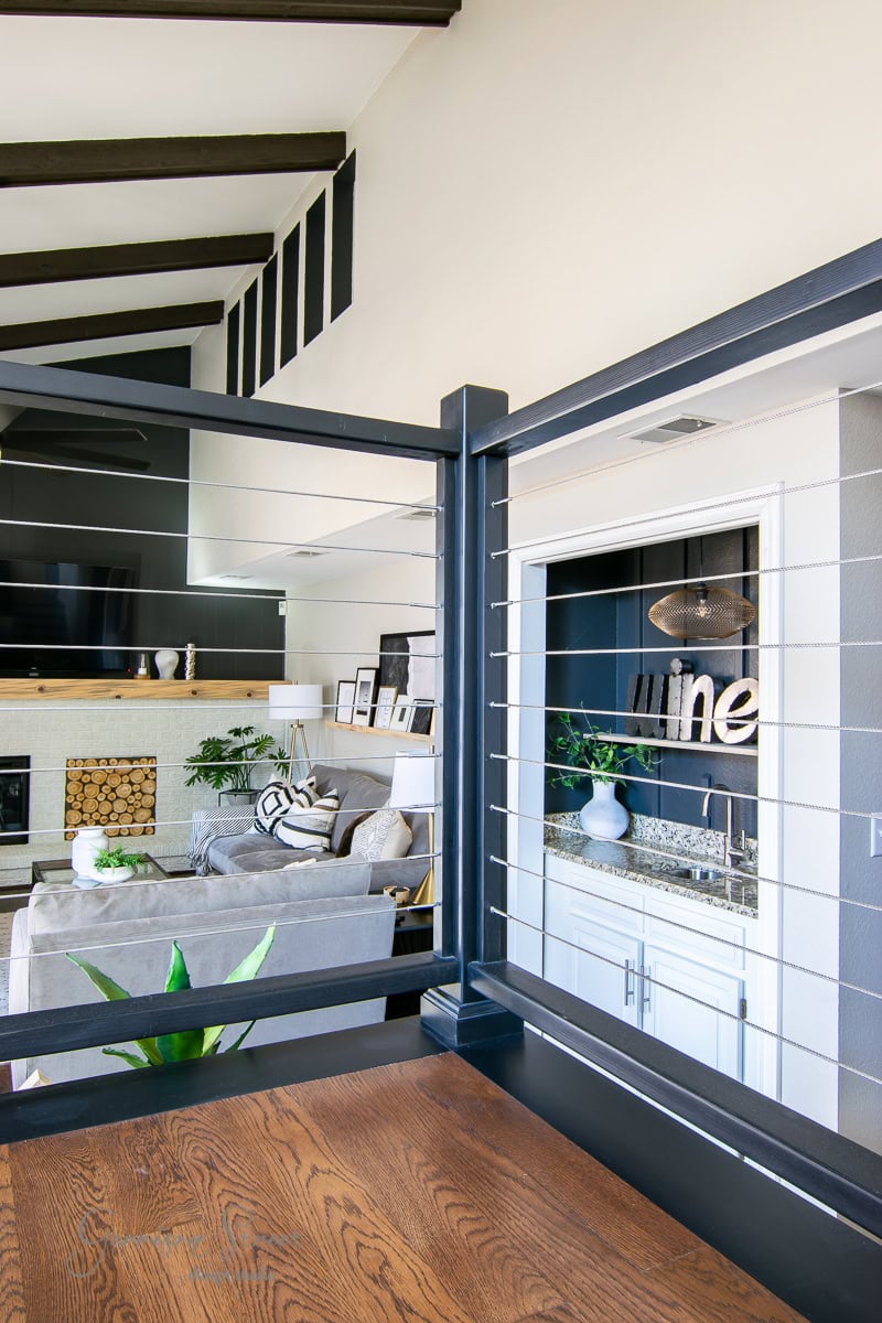 Modern living room with high ceiling and wooden beams. Features a cozy seating area with a gray couch, a fireplace with stacked wood, and a small bar area with "wine" sign. Wooden floors and sleek cable railing complete the contemporary design.