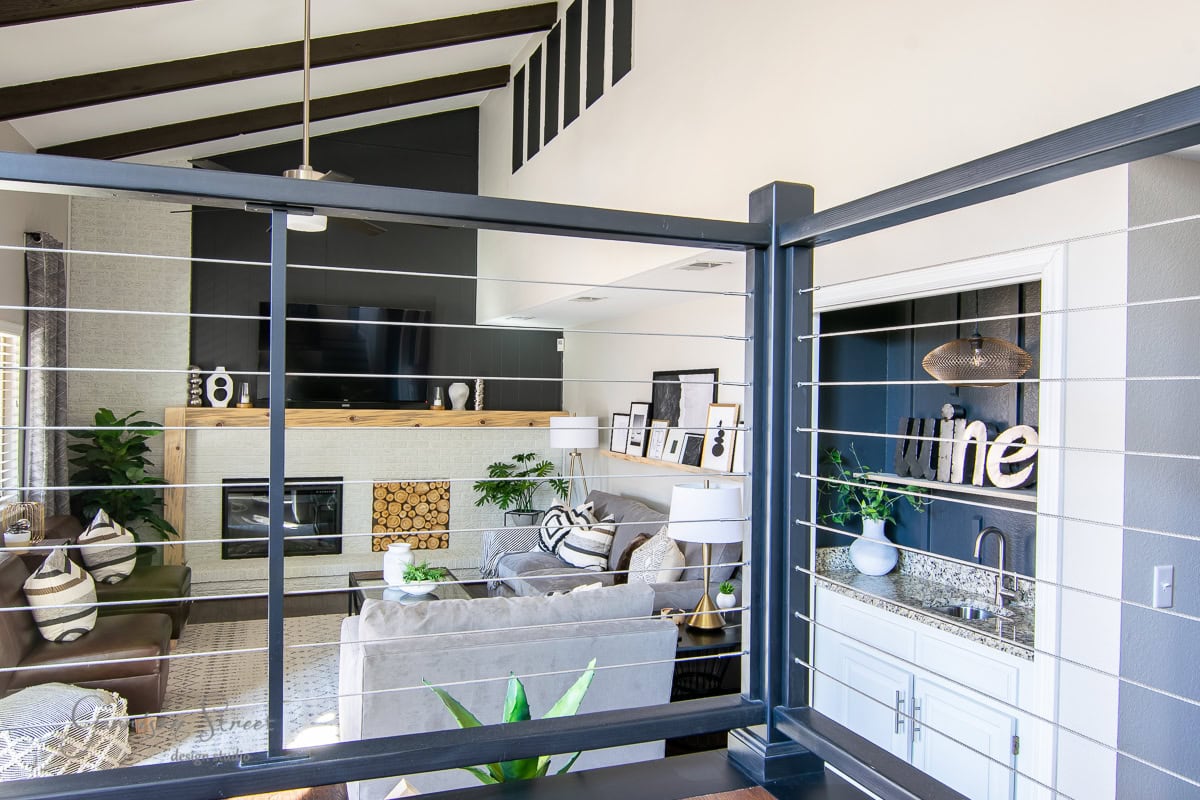 Spacious living room with high ceilings and exposed beams. Features a cozy seating area with a sectional sofa, large flat-screen TV, and fireplace. A wet bar with a "wine" sign is accented by DIY cable railing, while the decor includes plants and framed photos.