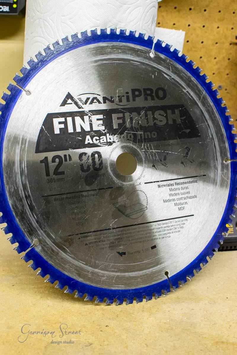 A 12-inch Avanti Pro fine finish saw blade with blue teeth and a metallic surface sits beside a roll of paper towels on a wooden workbench. Perfect for DIY enthusiasts, this blade is ideal for MDF projects, while the space is prepped for installing sleek cable railing systems.