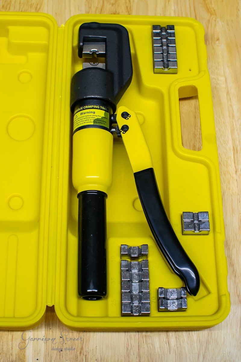 A yellow hydraulic hand crimping tool with a black handle rests in a matching yellow plastic case, perfect for any DIY cable railing project. The case neatly holds several metal dies around the tool, all set against a wooden surface backdrop.