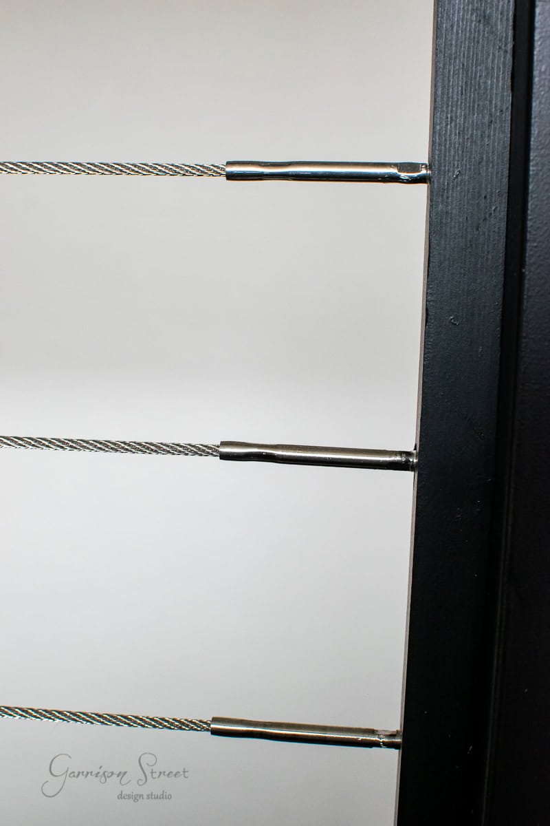 Close-up of a modern black railing featuring horizontal stainless steel cables. This sleek cable railing design is neatly aligned, showcasing a minimalist aesthetic. The plain background emphasizes its elegant lines, making it an ideal DIY project for contemporary spaces.