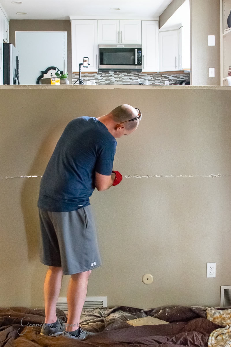 A person in casual clothes is engaged in a DIY project, using a tool to cut into drywall inside a home. The wall separates the kitchen in the background, featuring light cabinets and a colorful backsplash. A blanket covers the floor as they prepare for cable railing installation.