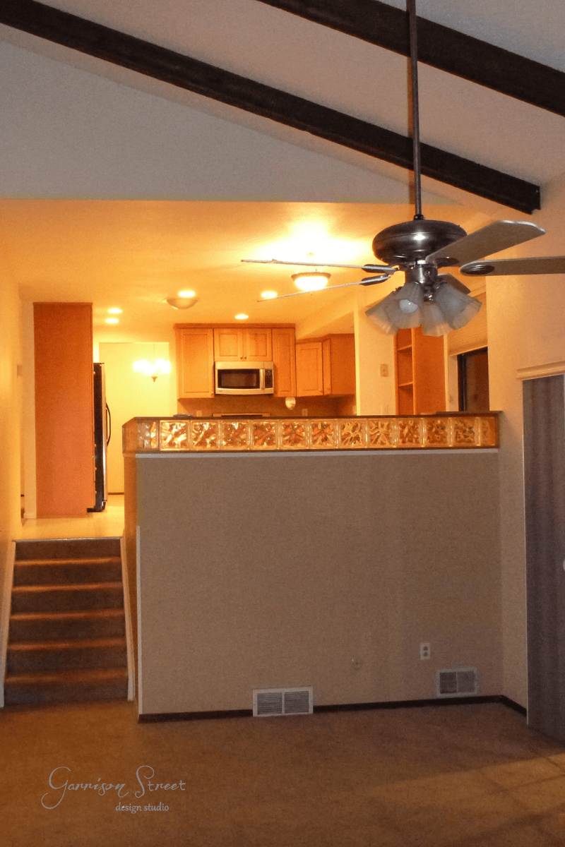 An interior view of a room with a high ceiling and exposed beams showcases a stylish DIY touch. A ceiling fan hangs overhead, while stairs with sleek cable railing lead up to a kitchen featuring wooden cabinets and a microwave. Glass tiles decorate the half wall, and warm lighting illuminates the space.