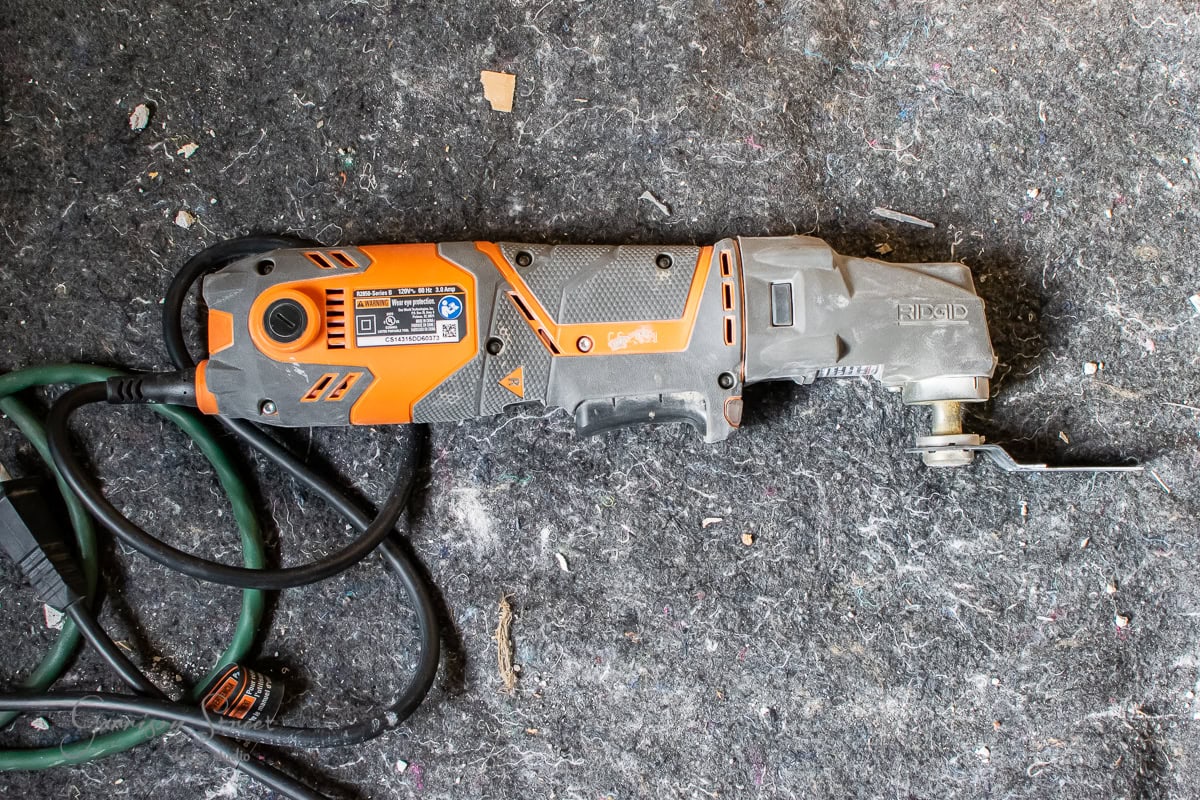 An orange and black multi-tool with a power cord rests on a textured gray surface, perfect for any DIY projects. The tool boasts a label featuring various symbols and brands, adding to its robust appeal.