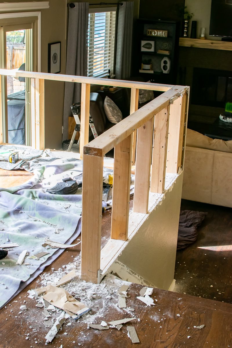 The living room is under a DIY renovation, featuring a partially dismantled wooden railing soon to be upgraded to sleek cable railing. Scattered debris and tools cover the floor, while windows and a fireplace are visible in the background, along with curtains and a couch.