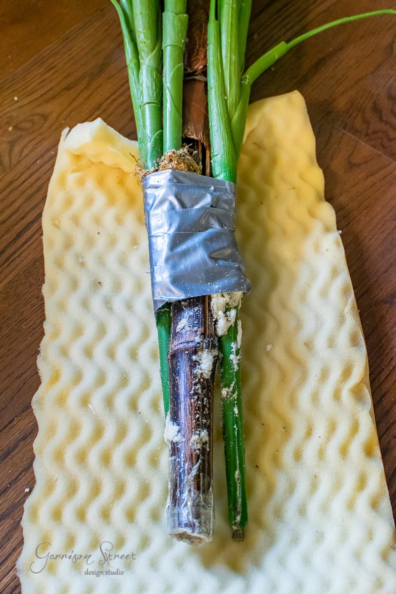 A DIY bundle of green plant stems wrapped in duct tape rests on a piece of crinkled white foam, resembling something made for a large planter.