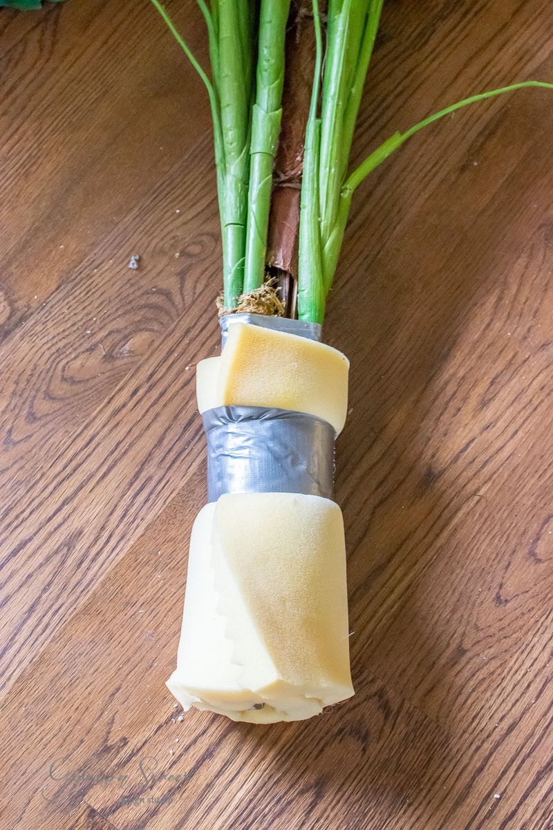 A DIY bundle of green stems wrapped with duct tape and surrounded by yellow foam padding lies on the wooden floor, waiting to be placed in the large planter.