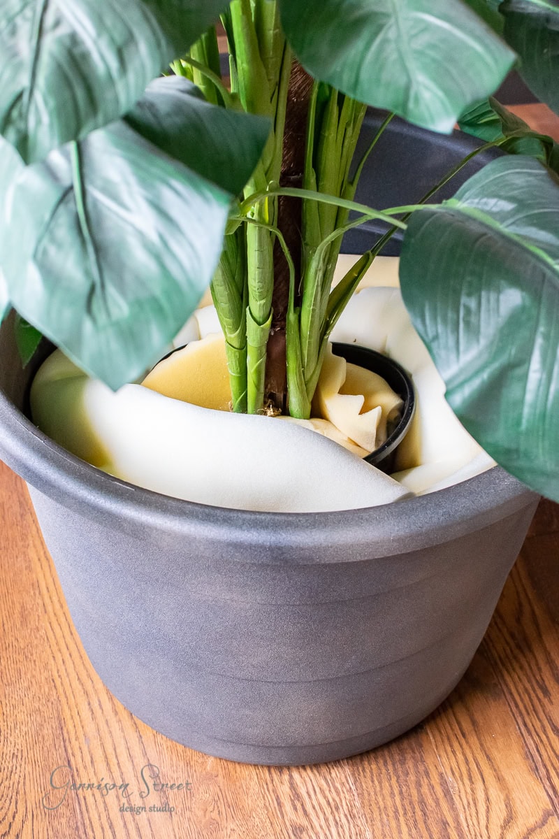 A potted plant with large green leaves is snuggly supported in a DIY gray pot using foam padding. The plant rests on a wooden floor, stabilized by the foam wrapped around its base, presenting an elegant alternative to a large planter.