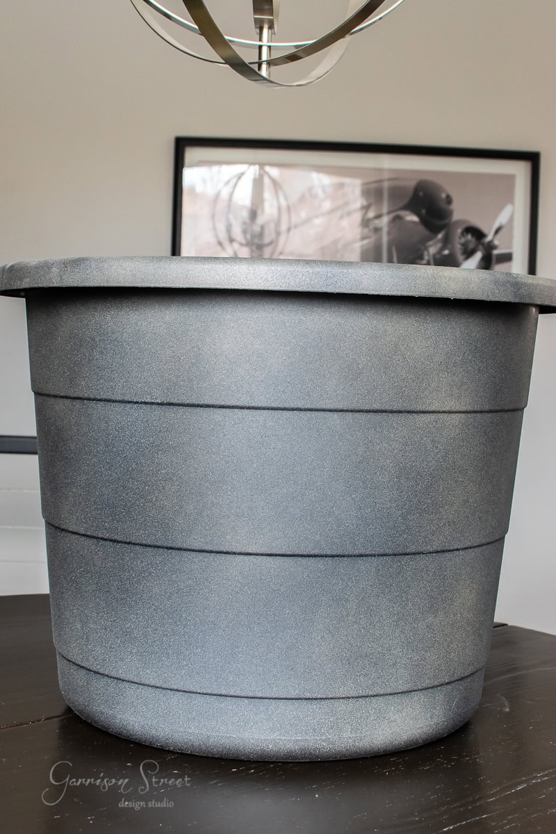 A large, round, gray planter with a textured finish sits on a dark wooden table, exuding DIY charm. In the background, there's modern art and a metallic light fixture above.