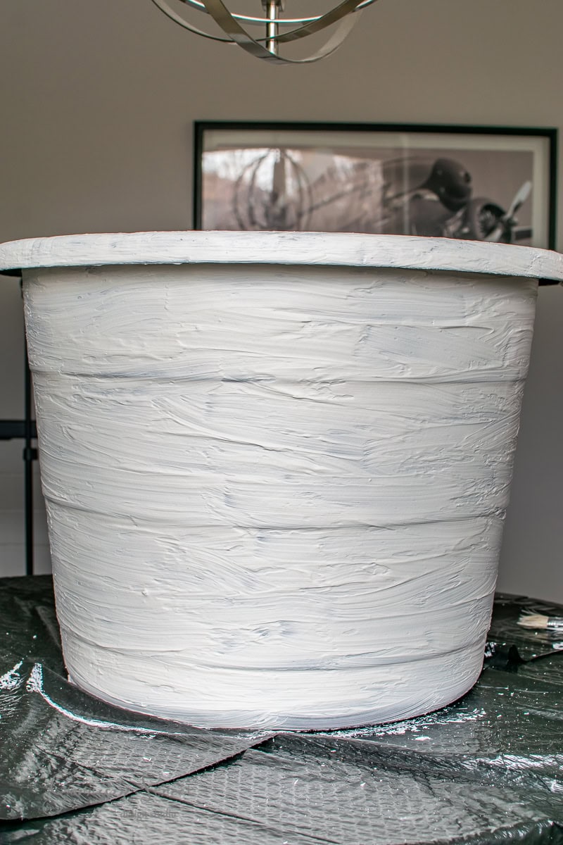 A large white textured pot, perfect as a DIY large planter, sits gracefully on a dark plastic sheet. In the background, a framed black and white photograph adds an artistic touch to the scene.