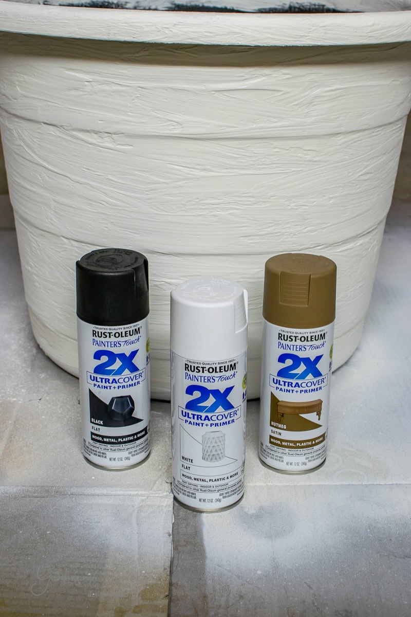 Three cans of Rust-Oleum Painter's Touch 2X Ultra Cover paint and primer are displayed in front of a large, white-painted planter. Perfect for DIY projects, the black, white, and brown cans provide amazing coverage on materials like metal, wood, and plastic.
