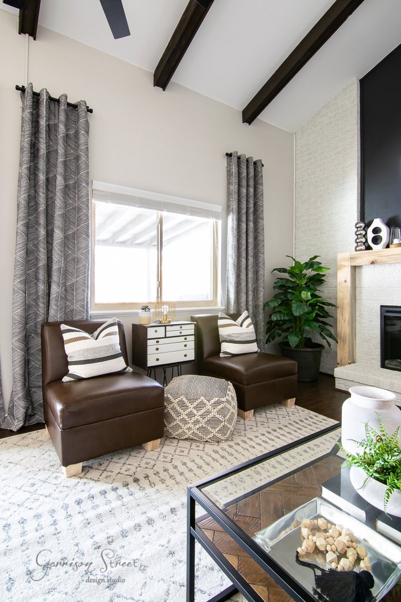 A modern living room with a high ceiling features two brown leather chairs, a geometric-patterned ottoman, and a small white cabinet. A DIY large planter hosts a towering plant in the corner. The space is beautifully accented with gray curtains and a patterned rug.