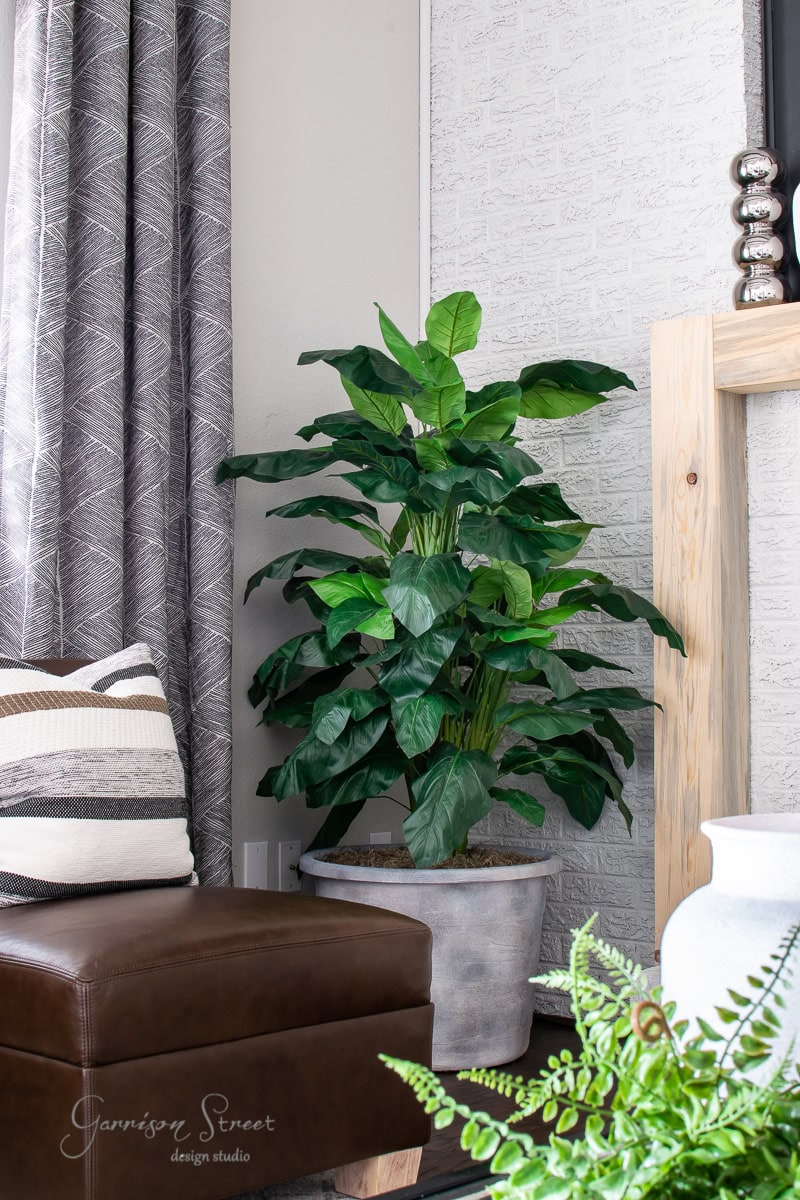 A lush green potted plant in a large planter sits on the floor near a light gray brick wall, next to a leather chair with striped cushions. A patterned curtain hangs in the background, adding texture to the cozy, modern living space. Consider a DIY touch for added personalization.