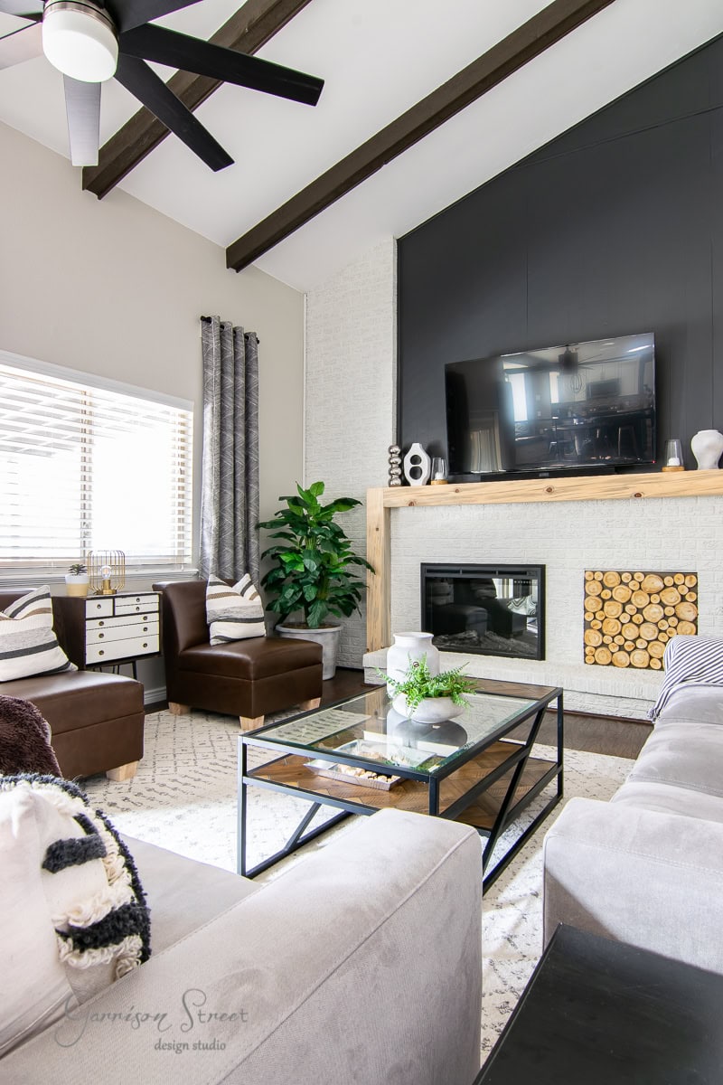 A modern living room with a high ceiling and exposed beams features a wall-mounted TV above the fireplace. The space includes a gray sectional sofa, brown armchairs, and a glass coffee table alongside a DIY touch with logs in the fireplace opening and a large planter housing a lush potted plant.