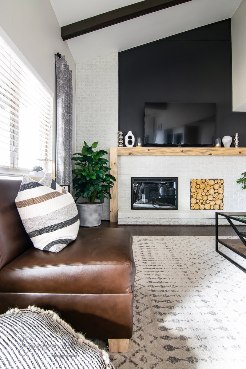 Modern living room with a brown leather sofa, striped pillow, and textured rug. A sleek fireplace with stacked wood accents is centered under a mounted TV. Large planters flank the fireplace, while natural light filters through the window blinds, adding a DIY touch of greenery.