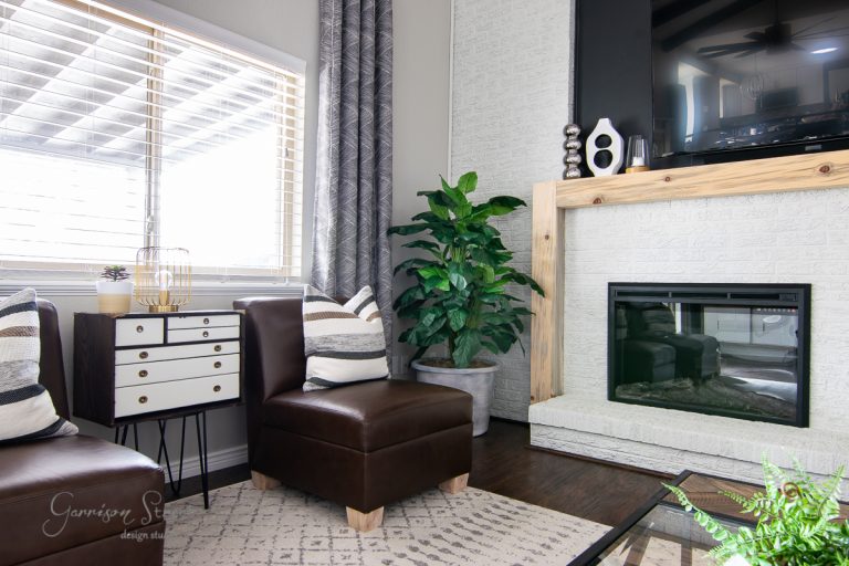 A modern living room features a brown leather chair, small white cabinet, and decorative pillows. A large planter sits in the corner next to the DIY fireplace with a wooden mantle and wall-mounted TV above it. A window with blinds lets in natural light.