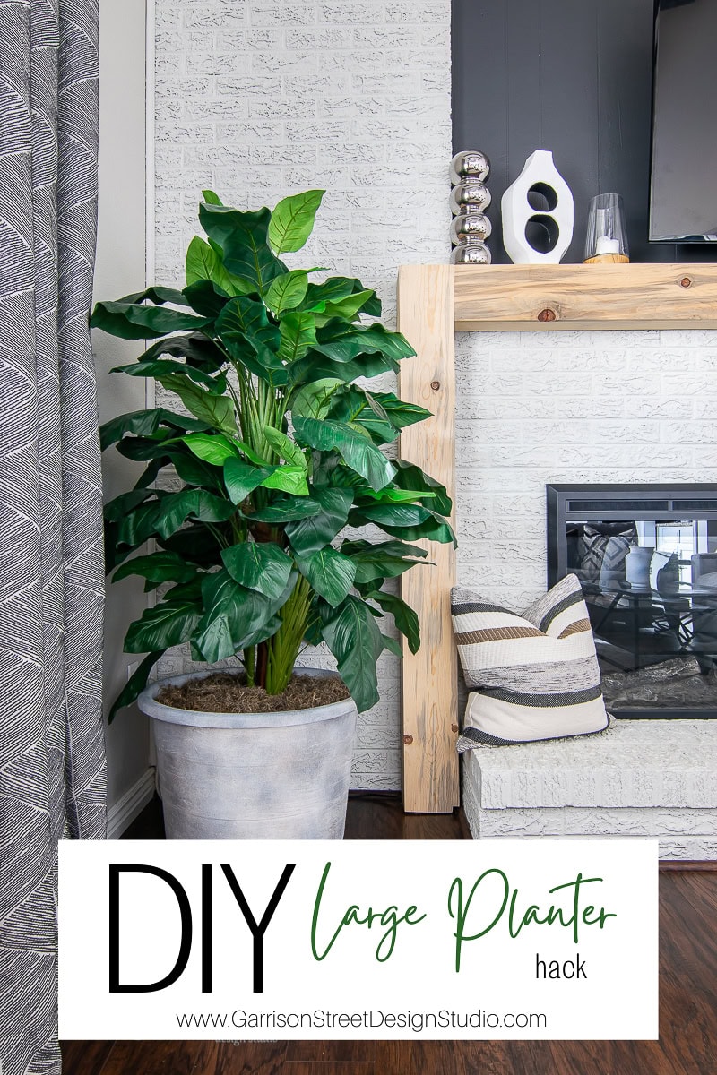 A cozy living room showcases a DIY large planter with a vibrant green plant beside the fireplace. Decorative items adorn the mantel, and a TV is mounted above. A patterned curtain is partially drawn to the side, adding charm to this inviting space. Text on the image reads “Large Planter Hack.”.