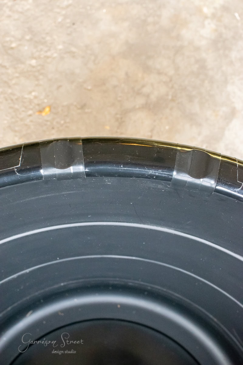 Close-up view of a sleek black plastic or metal container with several horizontal ridges, reminiscent of a large planter perfect for DIY projects. The smooth, shiny surface contrasts against the textured, concrete-like background.
