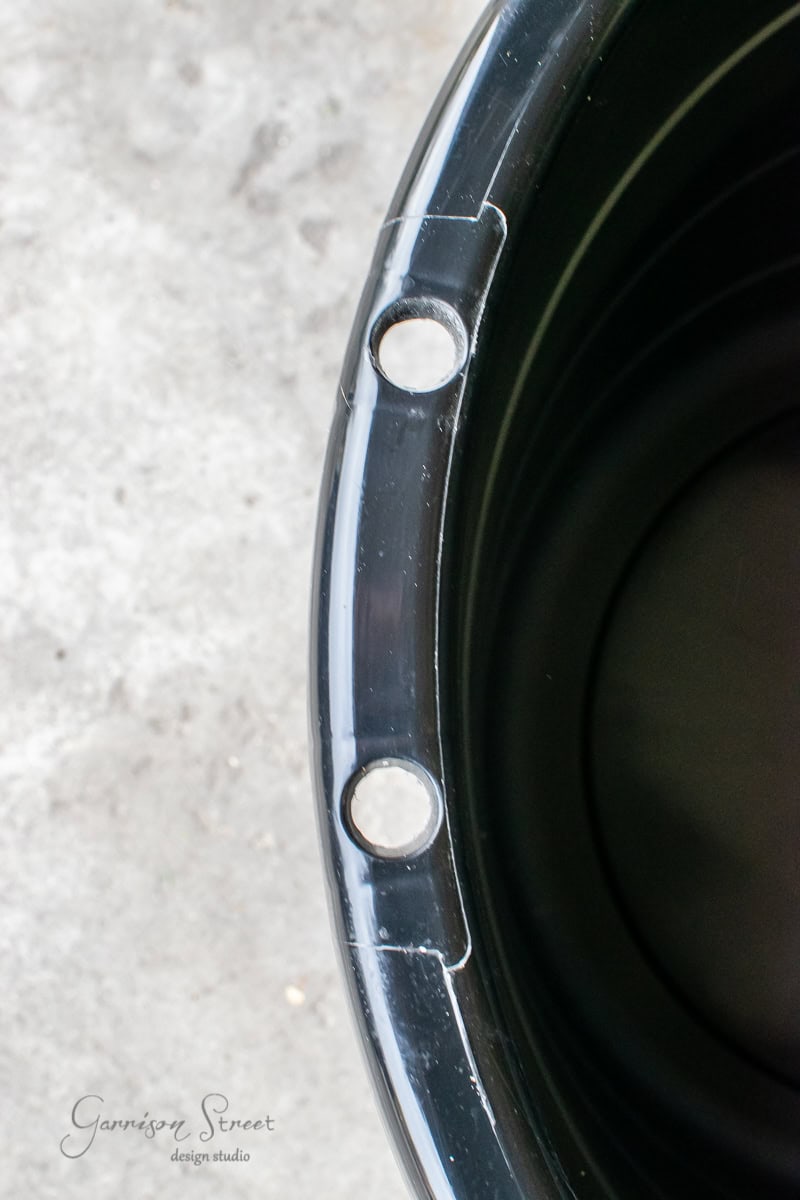 Close-up of a black plastic bin, repurposed as a large planter, with two holes near the rim on a concrete surface. The text "Garrison Street design studio" is visible on the lower left corner, showcasing a touch of DIY creativity.