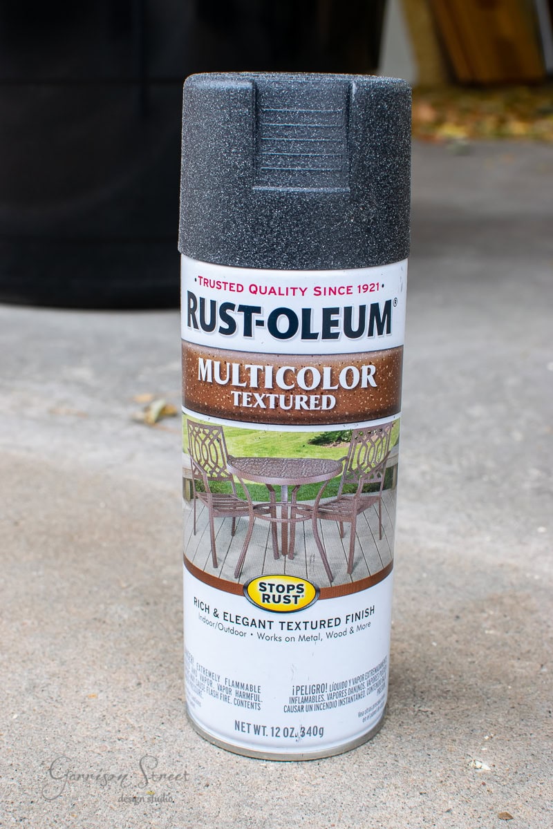 A can of Rust-Oleum multicolor textured spray paint is standing on a concrete surface, perfect for your DIY projects. The label shows a painted outdoor table and chairs, and the can promises a rich, elegant textured finish that stops rust—ideal for revitalizing that large planter.