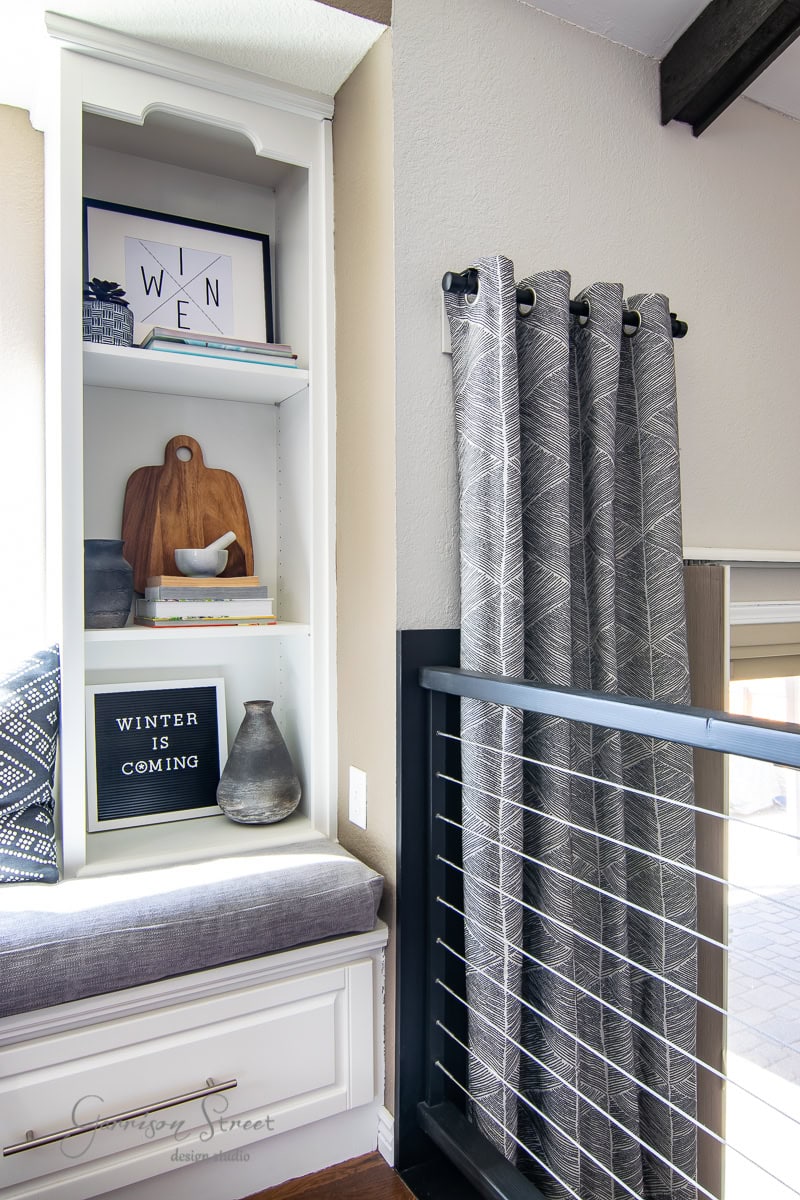 A cozy corner with built-in white shelves displaying books, a wooden cutting board, an IKEA vase, and a decor sign saying "Winter is Coming." The window seat has patterned cushions supported by a DIY metal railing, with gray curtains elegantly hanging from a sleek curtain rod.
