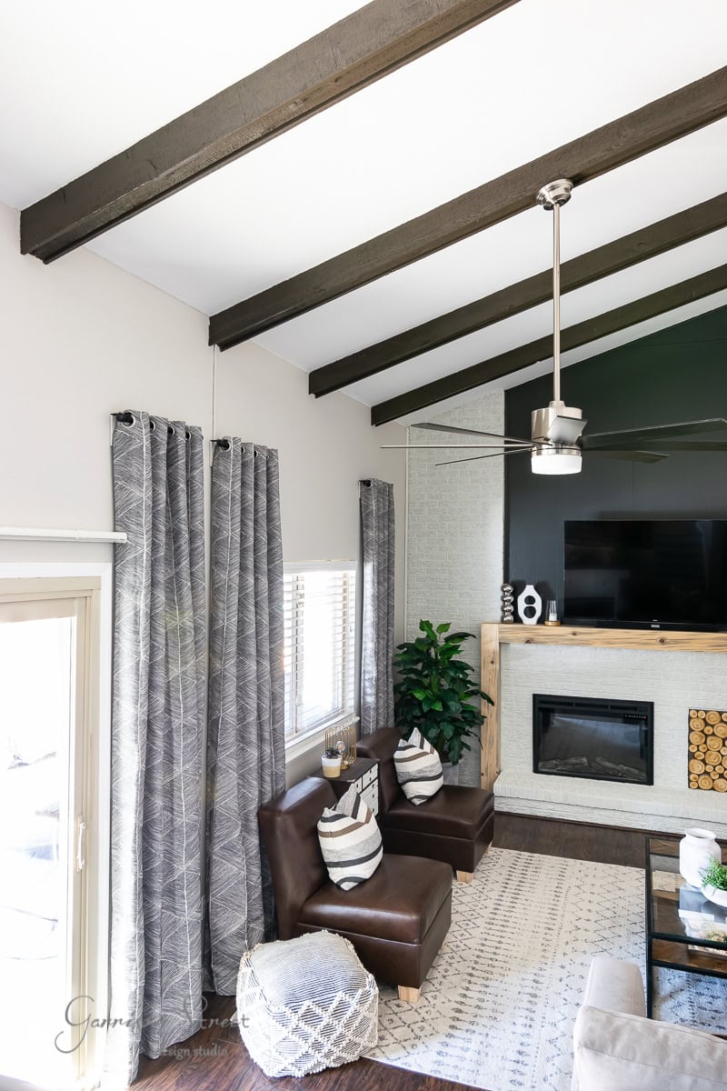 A cozy living room with high, beamed ceilings features a modern fireplace with wood stored underneath, gray patterned IKEA curtains hanging from a sleek curtain rod, brown leather chairs, a geometric pouf, and potted plants. A ceiling fan gently spins above a white and beige rug.