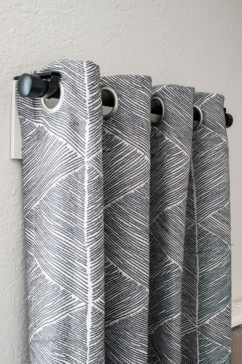 Close-up of hanging curtains with a geometric pattern in black and white. The design features a series of parallel lines creating a symmetrical, zigzag look. Threaded through a curtain rod, they complement the light gray wall.