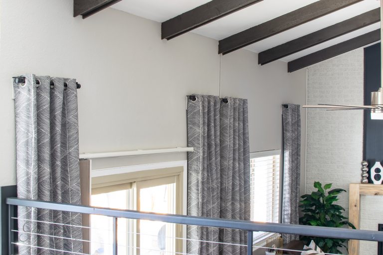A modern interior room with high ceilings and exposed wooden beams features large windows dressed in patterned gray curtains hanging from sleek IKEA curtain rods. A metal railing complements the potted plant and part of a ceiling fan, while neutral wall colors enhance the open space.