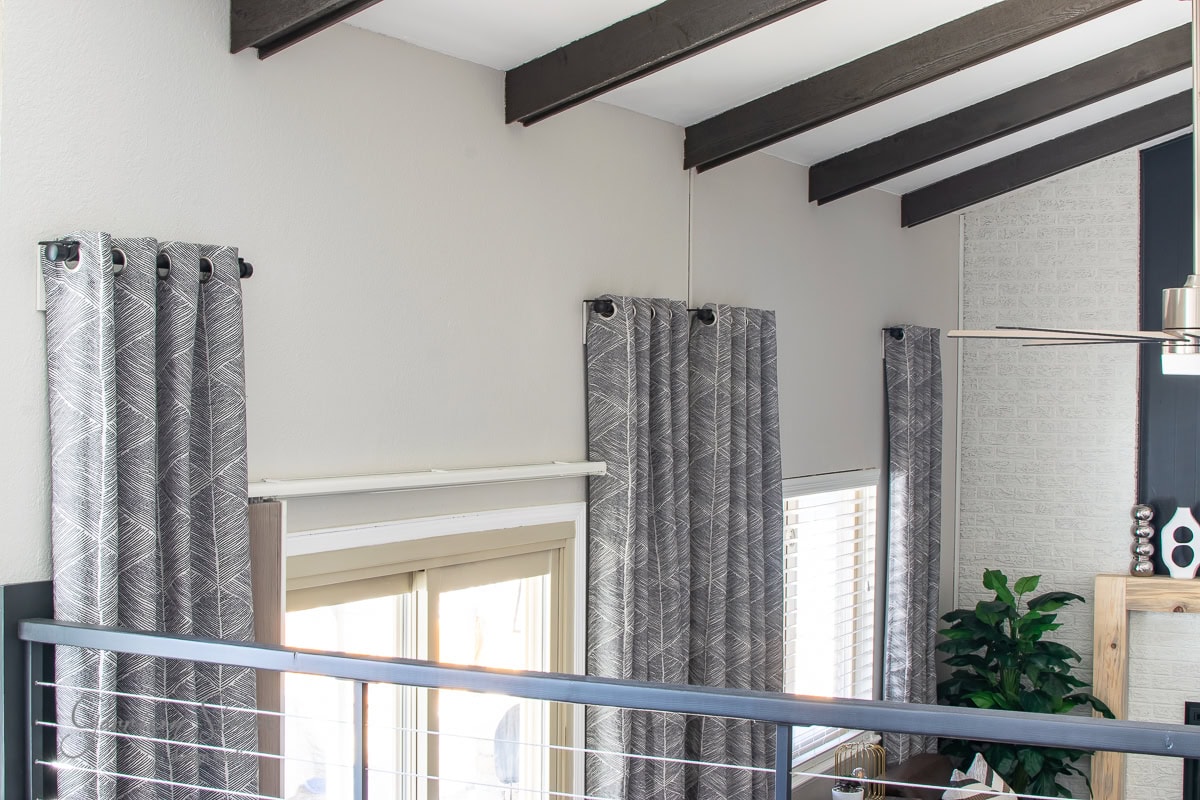 A modern interior room with high ceilings and exposed wooden beams features large windows dressed in patterned gray curtains hanging from sleek IKEA curtain rods. A metal railing complements the potted plant and part of a ceiling fan, while neutral wall colors enhance the open space.