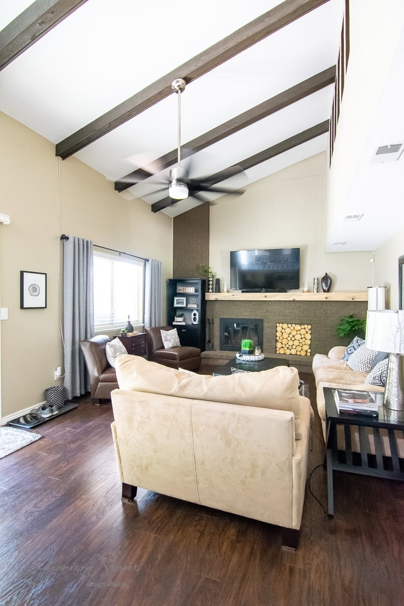 A cozy living room with high ceilings, exposed beams, and a ceiling fan. An IKEA beige sofa and armchairs face a flat-screen TV above the fireplace with stacked firewood. Wooden floors complement the large window, adorned with gray curtains hung on a sleek curtain rod.