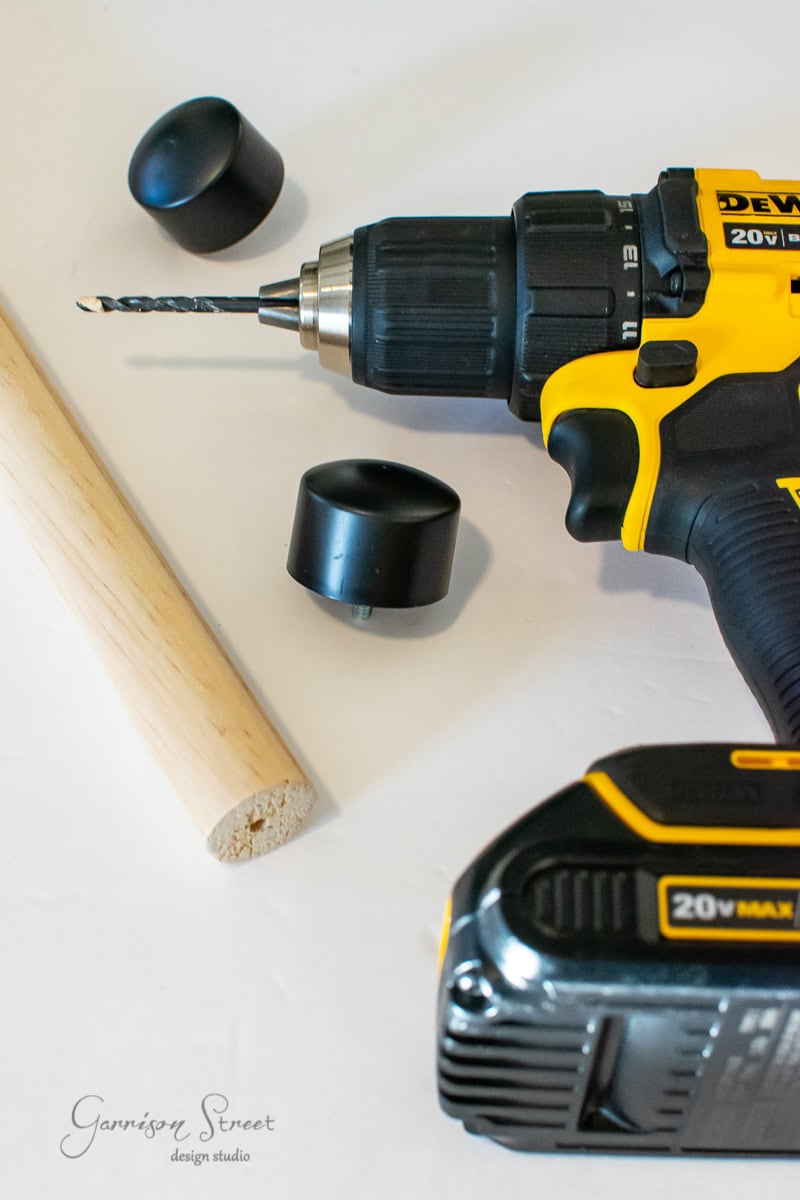 A yellow and black cordless power drill with a drill bit rests on a white surface. Nearby, you'll find two black rubber cane tips, a wooden dowel, and the makings of a DIY curtain rod project. The image includes a watermark reading "Garrison Street Design Studio."