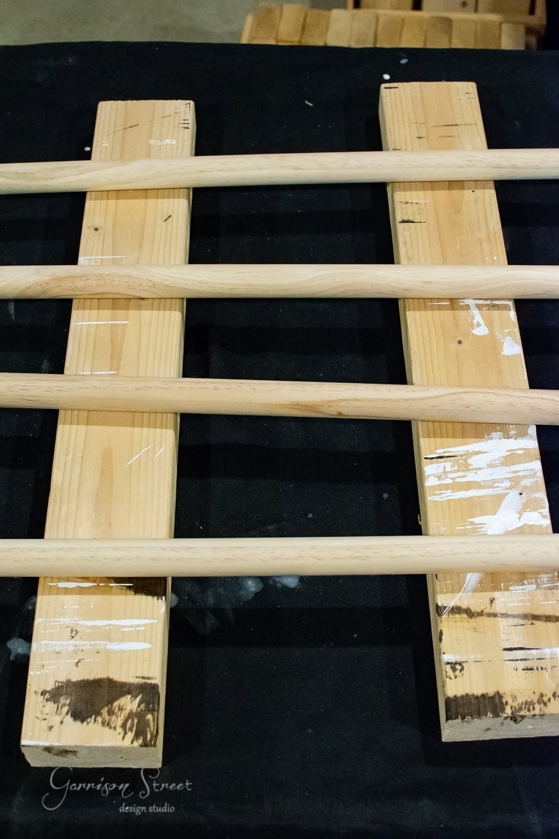 Four wooden rods rest horizontally over two parallel planks on a black background. The planks, with their white paint stains, have a DIY charm that adds character to the arrangement.