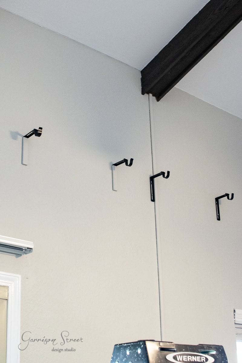 Four black wall brackets are mounted on a light gray wall, evenly spaced and waiting for a curtain rod. A beam runs across the ceiling while a part of a ladder is visible in the lower right corner, hinting at an ongoing DIY project. "Garrison Street Design Studio" is elegantly written on the wall.
