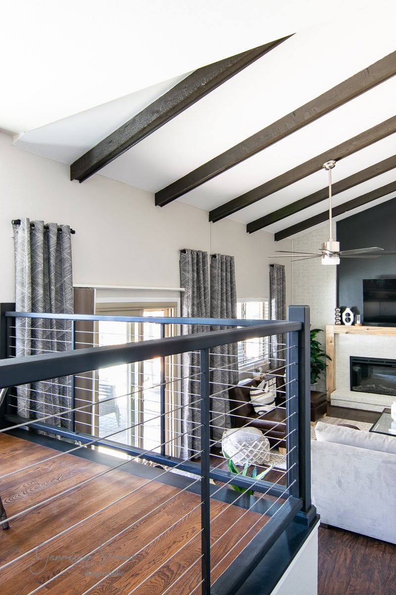 Modern living room with vaulted ceiling, exposed beams, and a metal railing. Hardwood floors and large windows featuring patterned curtains hung on a sleek curtain rod. A ceiling fan and flat-screen TV are visible, along with a cozy IKEA seating area and a fireplace.