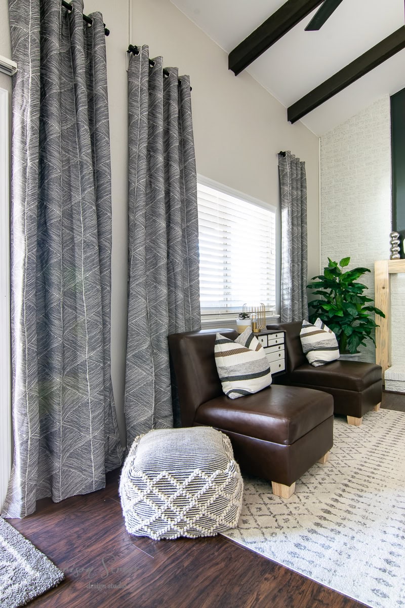 A cozy living space features brown leather chairs, patterned gray curtains elegantly hanging from a sleek curtain rod, and a white side table adorned with striped pillows. A textured pouf and a plant in the corner add comfort, while a rug enhances the dark wood floor.