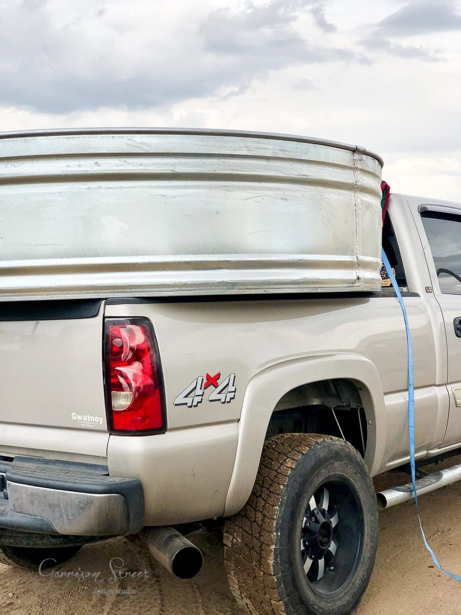 A silver pickup truck with a 4x4 logo has a large metal stock tank secured in its bed using a blue strap. The truck is parked on a dirt surface under a cloudy sky.