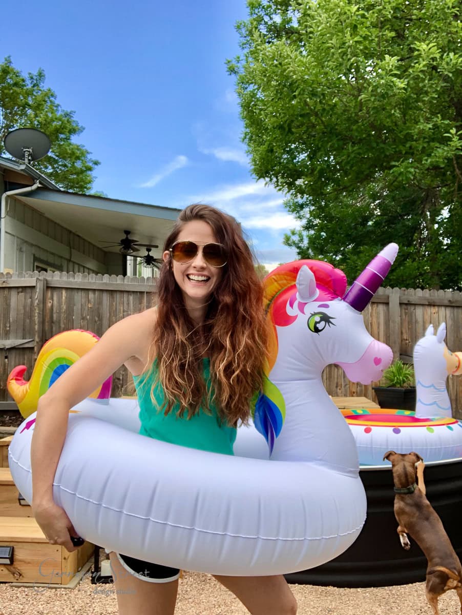 A woman with long brown hair and sunglasses smiles while holding a large inflatable unicorn pool float. She's standing in a backyard by a pool, with another inflatable and a small brown dog nearby. The sky is clear and blue.