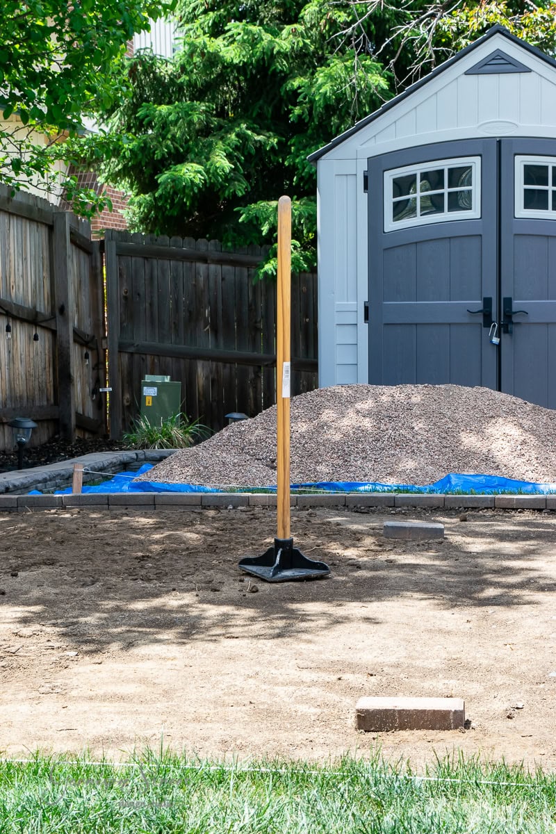 A backyard scene with a tamping tool standing upright on the ground. A blue tarp with a pile of gravel hints at preparations for a stock tank pool in front of a gray and white shed. Wooden fencing and green trees complete the picturesque setting.
