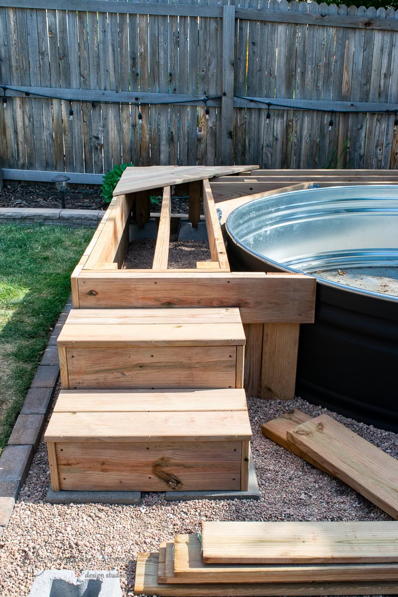 A partially constructed wooden deck surrounds an above-ground metal stock tank pool. Wooden steps lead up to the deck. The area is bordered by a wooden fence, and planks lie on the ground nearby, indicating ongoing construction.