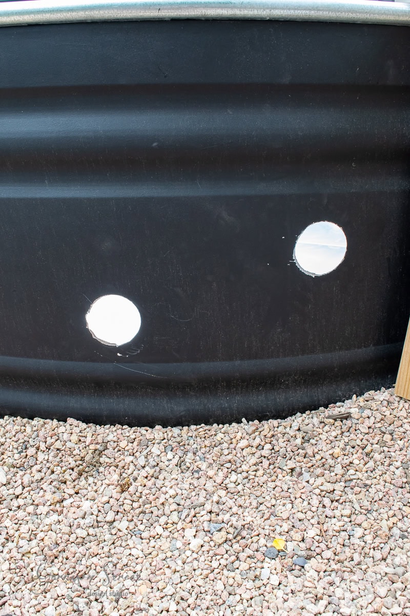 A black metal sheet with two circular holes is positioned above a bed of small gravel stones, hinting at the creation of a stock tank pool. Wooden objects are partially visible on the side, suggesting construction or repair work in progress.
