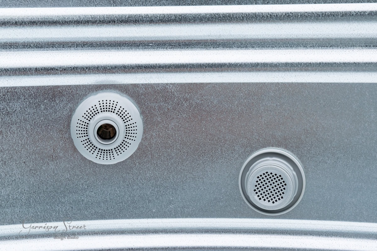 Close-up of a metal surface featuring two circular, perforated fixtures reminiscent of those on stock tanks. The background boasts a textured, metallic finish with horizontal grooves, evoking the rugged charm of poolside industrial design.